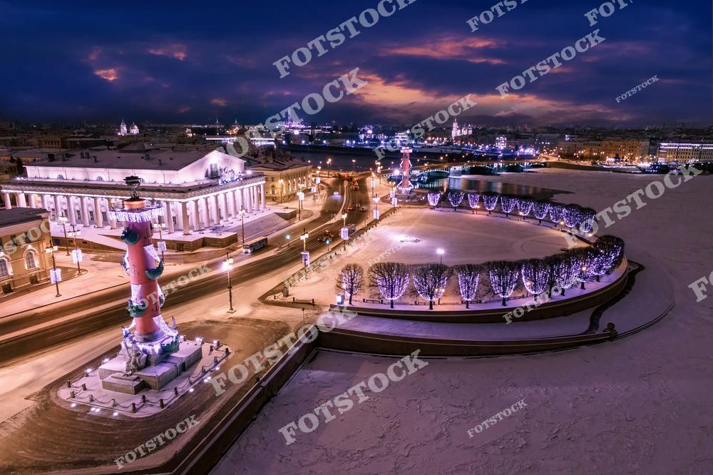 На фотографии изображена Стрелка Васильевского острова в зимний вечер. Ростральные колонны, украшенные гирляндами, выделяются на фоне ночного неба. Заснеженная площадь и улицы города сияют от огней, создавая праздничную атмосферу. Вдали видны исторические здания Санкт-Петербурга, а также река Нева, отражающая ночные огни. Этот вид с высоты квадрокоптера прекрасно передает атмосферу зимнего Петербурга в новогодние праздники.